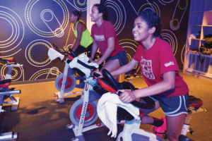 First-year student Lelandra Randle (right) attends her first spinning class with her floormates ­Millyn ­Brieschke Robertson (center) and ­Tenille Tubbs. Their instructor, Meghann Feely, assistant director of fitness and wellness, says she loves helping students develop healthy habits early. (Dan Donovan/Washington University)