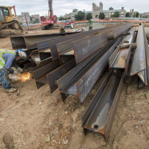 a man in a yellow vest and hard hat welds one of many metal beams in a tall pile. Sparks fly.