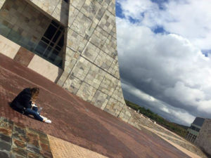 In a slanted photo, a girl sits on the floor next to a curved facade of a building, sketching in a book.