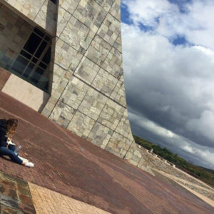 In a slanted photo, a girl sits on the floor next to a curved facade of a building, sketching in a book.