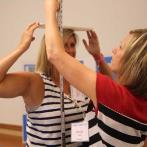 two women hold measuring sticks vertically and horizontally against their heads