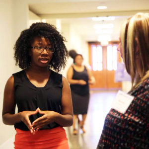 a young black woman with glasses presents a poster to a blonde judge in a bright hallway