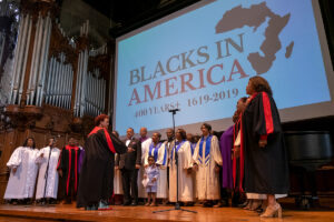 Gospel choir stands while singing on stage with Blacks in America slogan projected behind.