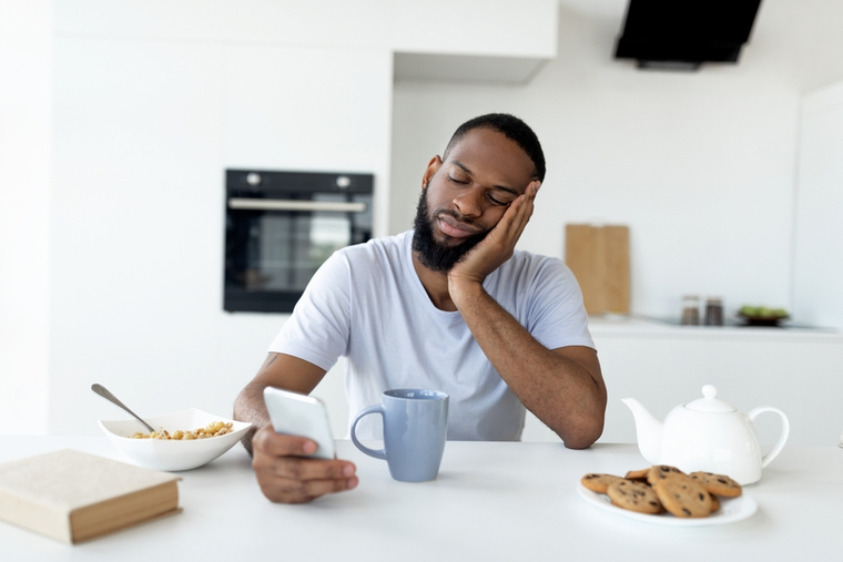 Man sitting at a table, doomscrolling. He's looking at his phone, looking depressed