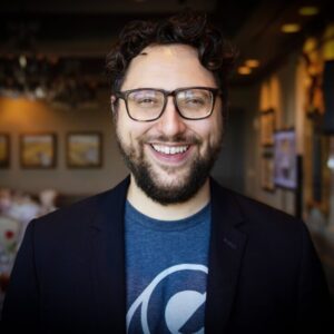 David Karandish, founder of Capacity, wearing a blue t-shrt and black jacket and smiling broadly.