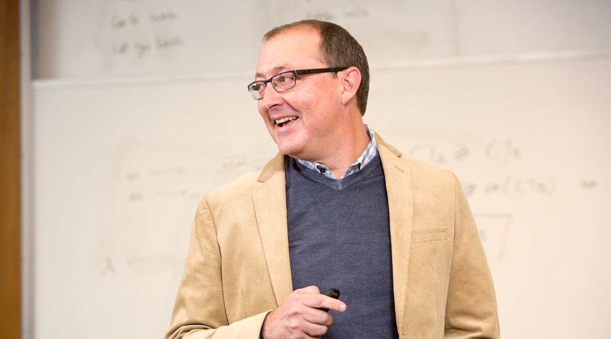 Man smiling in front of a whiteboard holding a black object Professor Doug Villhard