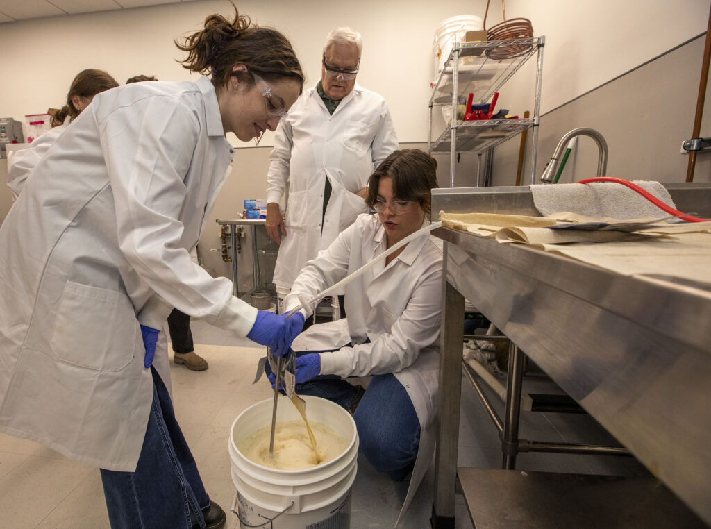 Two students pour liquid into a bucket while their professor looks on.