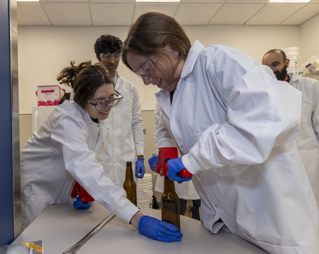 A student in a lab coat pushes down on a cap to finish sealing a bottle of beer.