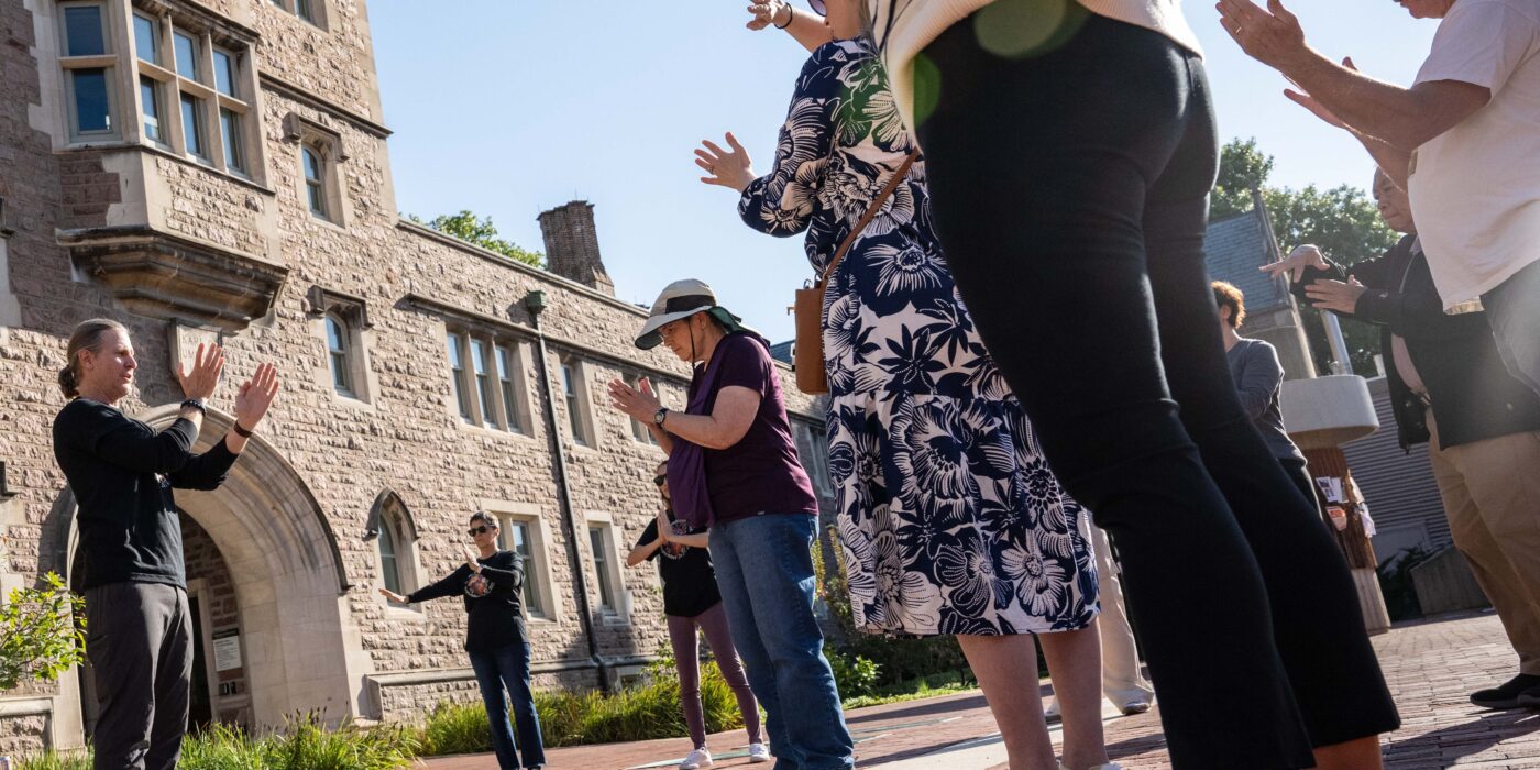 A group of people standing on a brick plaza hold hands up as a they do mindfulness movement at a university public space