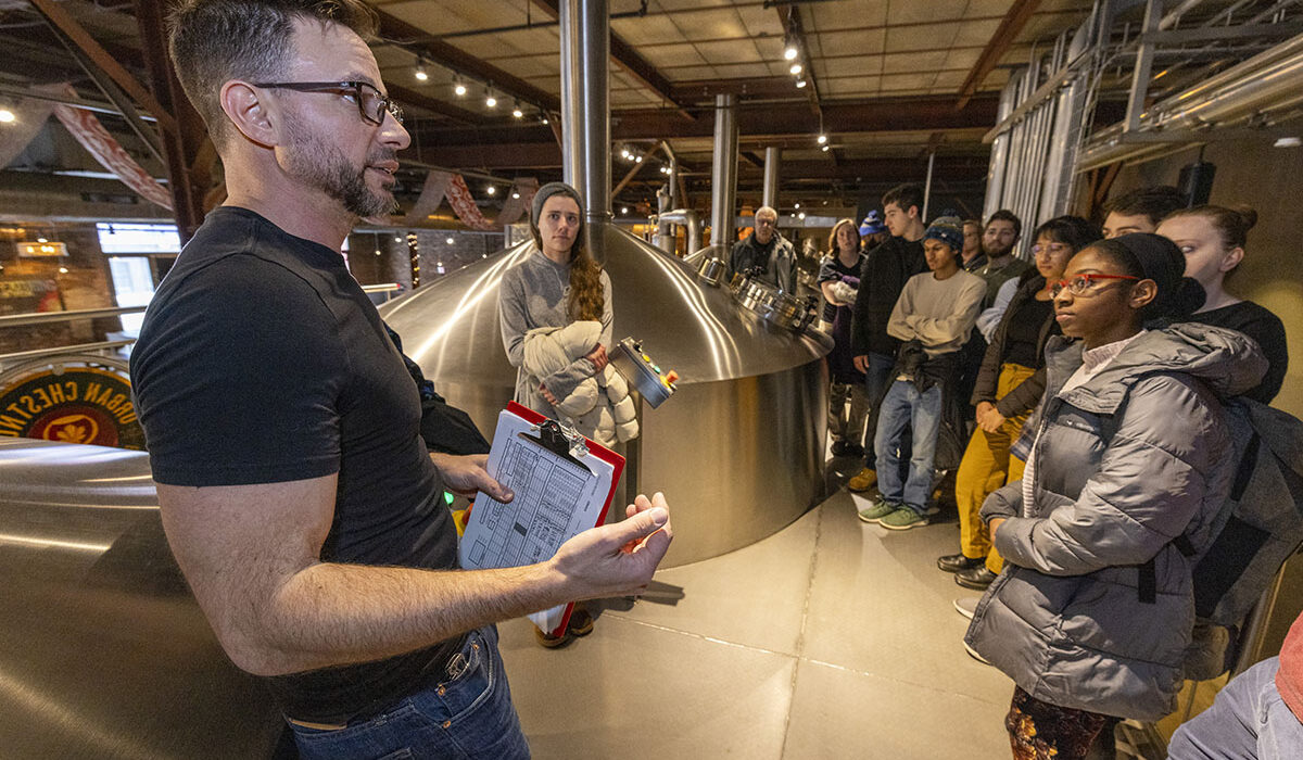 A man stands next to a brewing tank or bioreactor while students look on