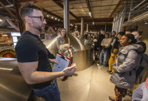 A man stands next to a brewing tank or bioreactor while students look on