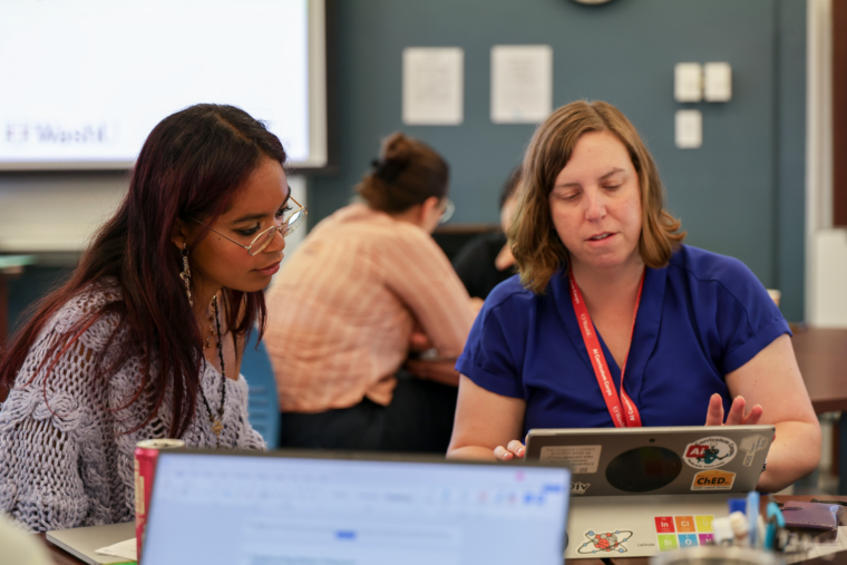 Two women sit at a small table and look into an open laptop. computer.