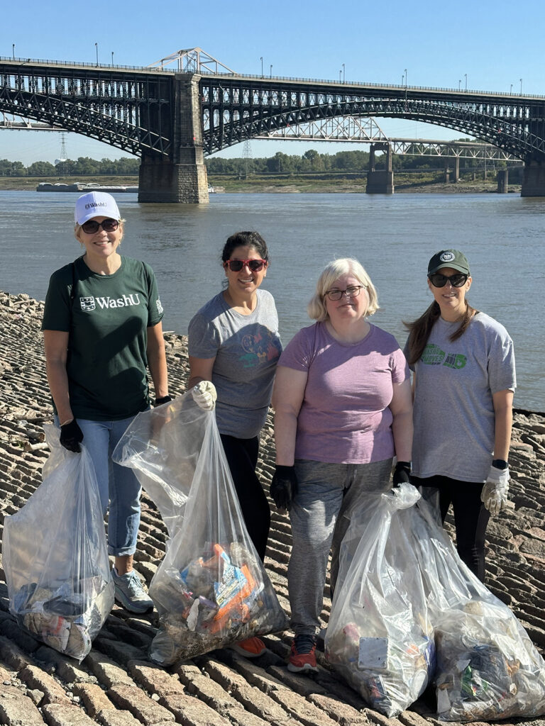 A group of volunteers stand with trash bags on the St. Louis riverfront with the Mississippi River in the background.