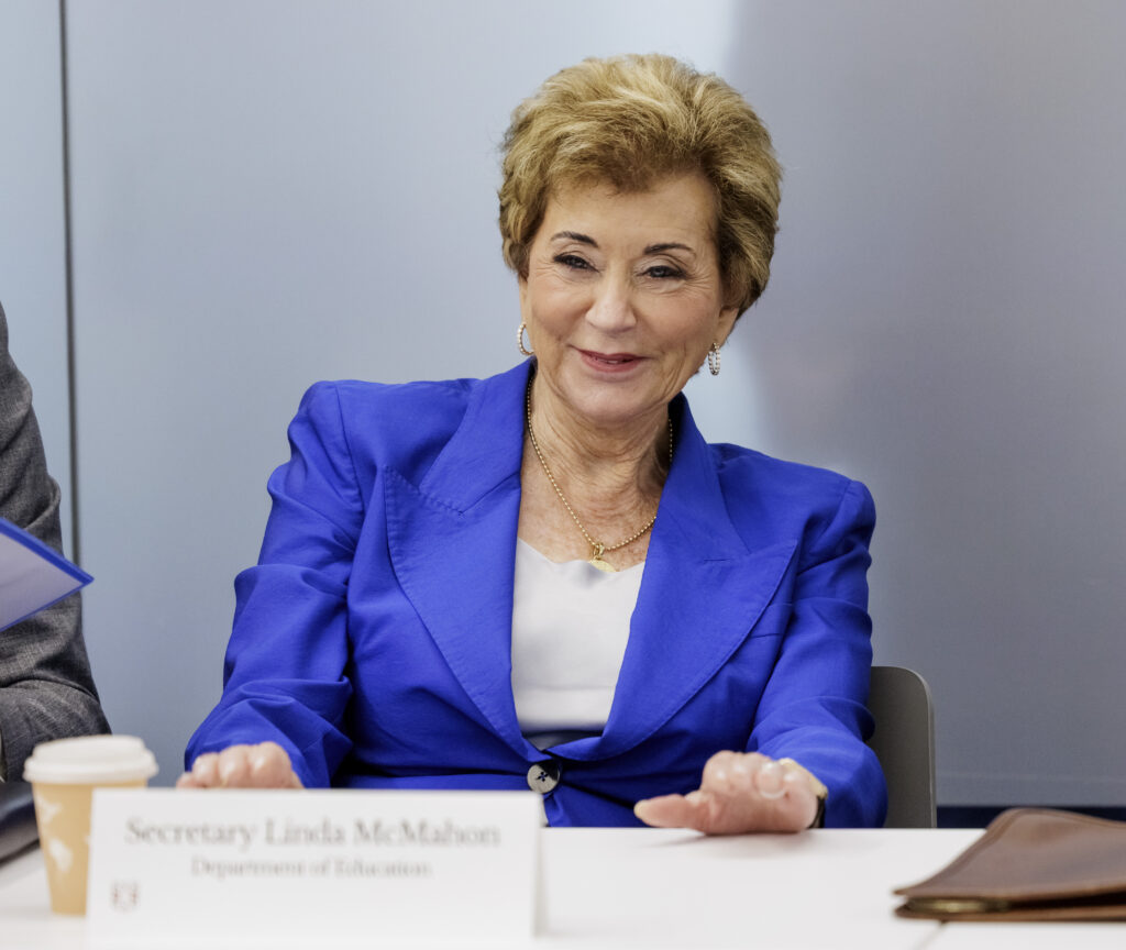 U.S. Education Secretary Linda McMahon sits in a chair at a conference table. She has short blond hair and is wearing a violet-colored suit jacket.