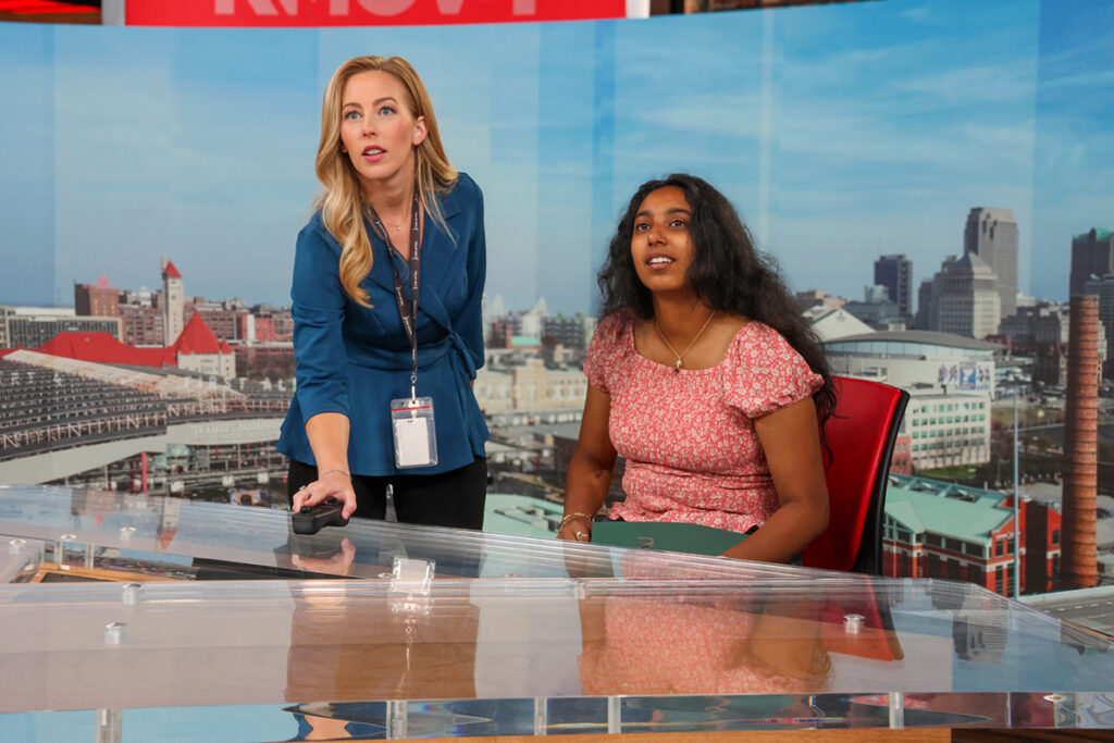 A student sits at the anchor desk of local TV station while the anchor looks on.